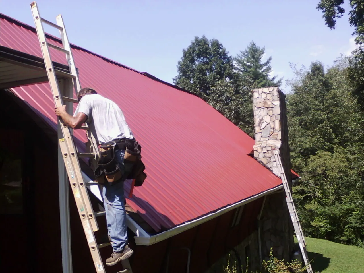 Roofer on a ladder beside a red metal roof and stone chimney — Soffit & Fascia Repair by Roofing Brothers in Lathrop